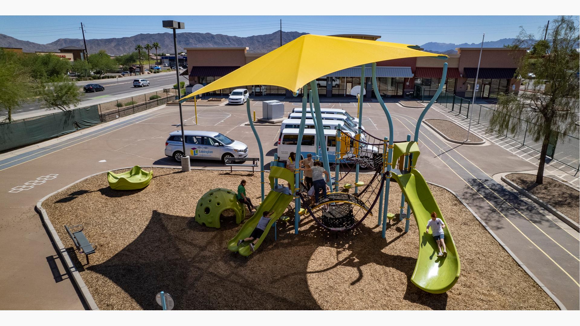 Elevated view of a children playing on an underwater themed play structure with a large yellow shade cover the entire structure. The play area and parking area filled with white vans sits in the middle of a running track.