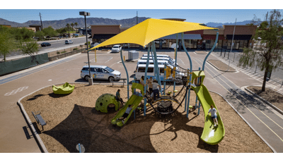 Elevated view of a children playing on an underwater themed play structure with a large yellow shade cover the entire structure. The play area and parking area filled with white vans sits in the middle of a running track.