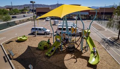 Elevated view of a children playing on an underwater themed play structure with a large yellow shade cover the entire structure. The play area and parking area filled with white vans sits in the middle of a running track.