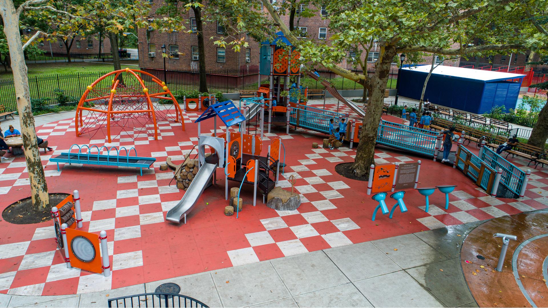 Elevated view of a city playground with a large inclusive play structure and a smaller play structure for younger children in the foreground. Grey and orange square safety surfacing create a checker pattern on the ground.