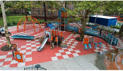 Elevated view of a city playground with a large inclusive play structure and a smaller play structure for younger children in the foreground. Grey and orange square safety surfacing create a checker pattern on the ground.