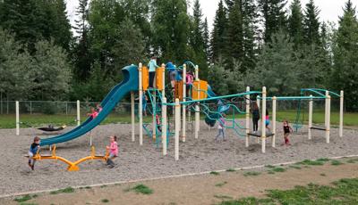 Blue and tangerine playground with many overhead items to play on. Kids playing on see-saw and going down slide. Many kids playing all around the playground. Tall green trees in the background with grey pebbles under the playground structure.