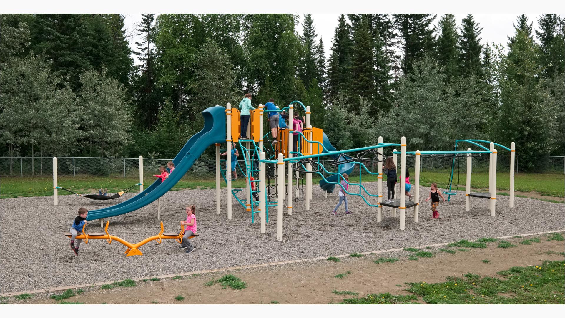 Blue and tangerine playground with many overhead items to play on. Kids playing on see-saw and going down slide. Many kids playing all around the playground. Tall green trees in the background with grey pebbles under the playground structure.