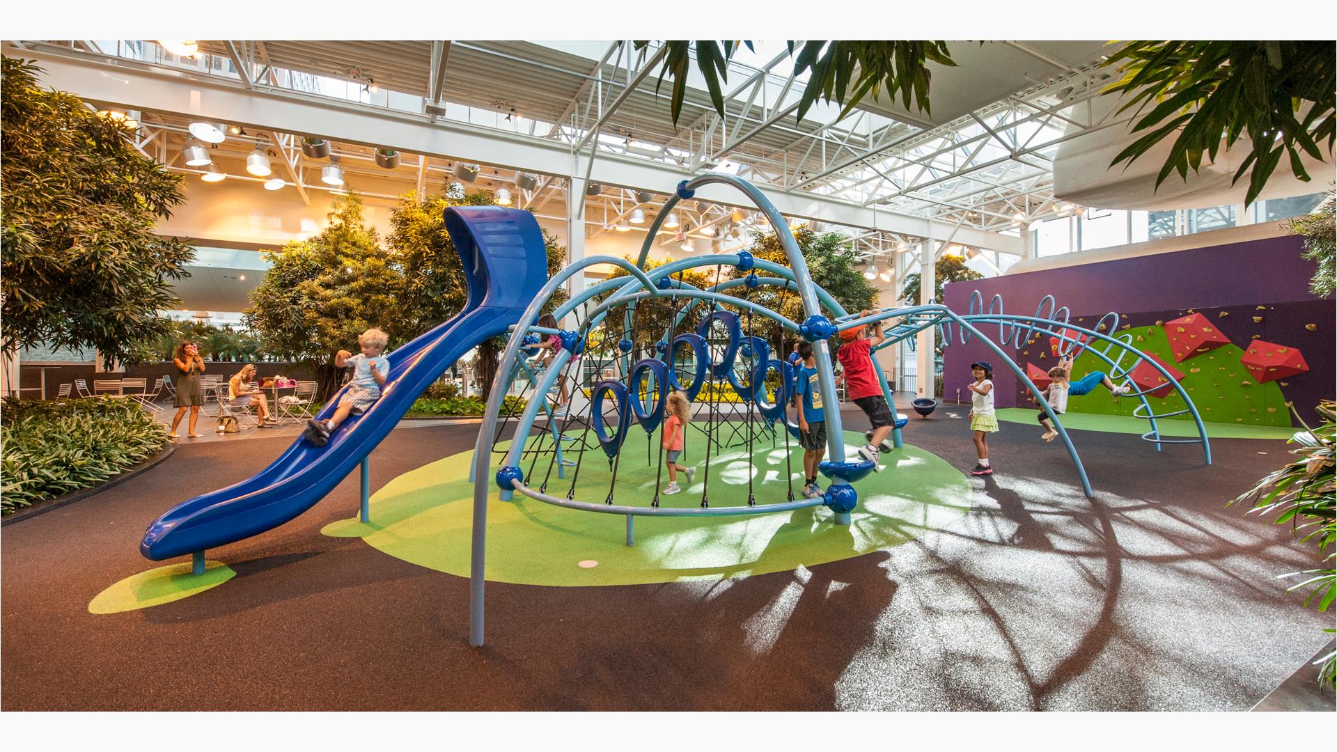 Inside a shopping center in Calgary, moms near some outdoor furniture take photos of their kids playing on this Evos play structure. The structure is surrounded by trees and long grass brush. A rock wall sits in the the background.