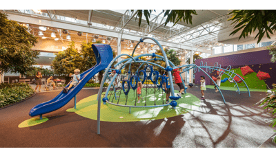 Inside a shopping center in Calgary, moms near some outdoor furniture take photos of their kids playing on this Evos play structure. The structure is surrounded by trees and long grass brush. A rock wall sits in the the background.