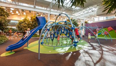 Inside a shopping center in Calgary, moms near some outdoor furniture take photos of their kids playing on this Evos play structure. The structure is surrounded by trees and long grass brush. A rock wall sits in the the background.