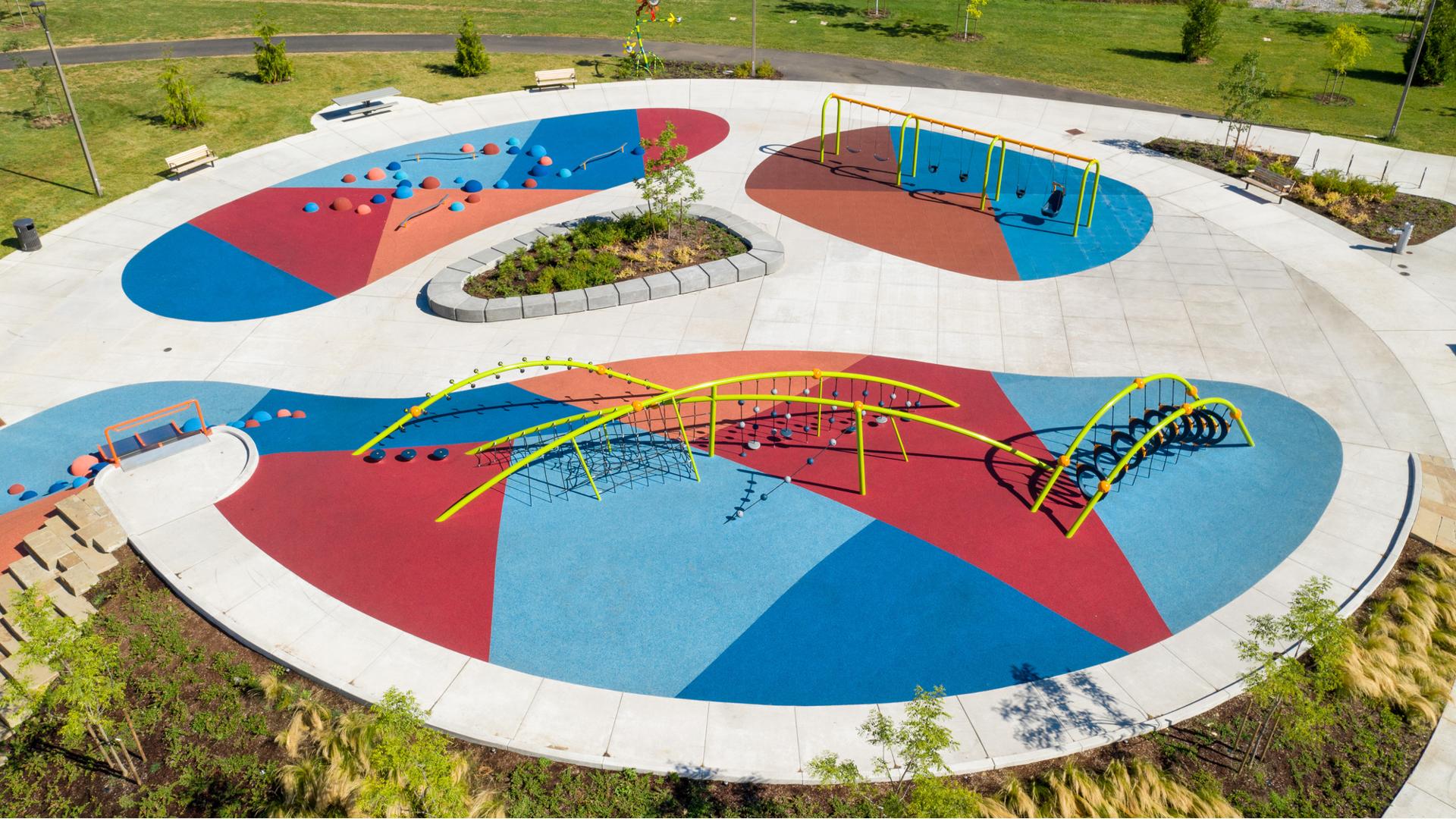 Aerial view of playground space with bright green net climbers with arched shaped posts. Play space also includes climbing pods and swings with blue, red and orange rubber surfacing. 