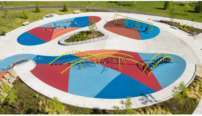 Aerial view of playground space with bright green net climbers with arched shaped posts. Play space also includes climbing pods and swings with blue, red and orange rubber surfacing. 