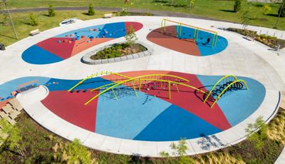 Aerial view of playground space with bright green net climbers with arched shaped posts. Play space also includes climbing pods and swings with blue, red and orange rubber surfacing. 