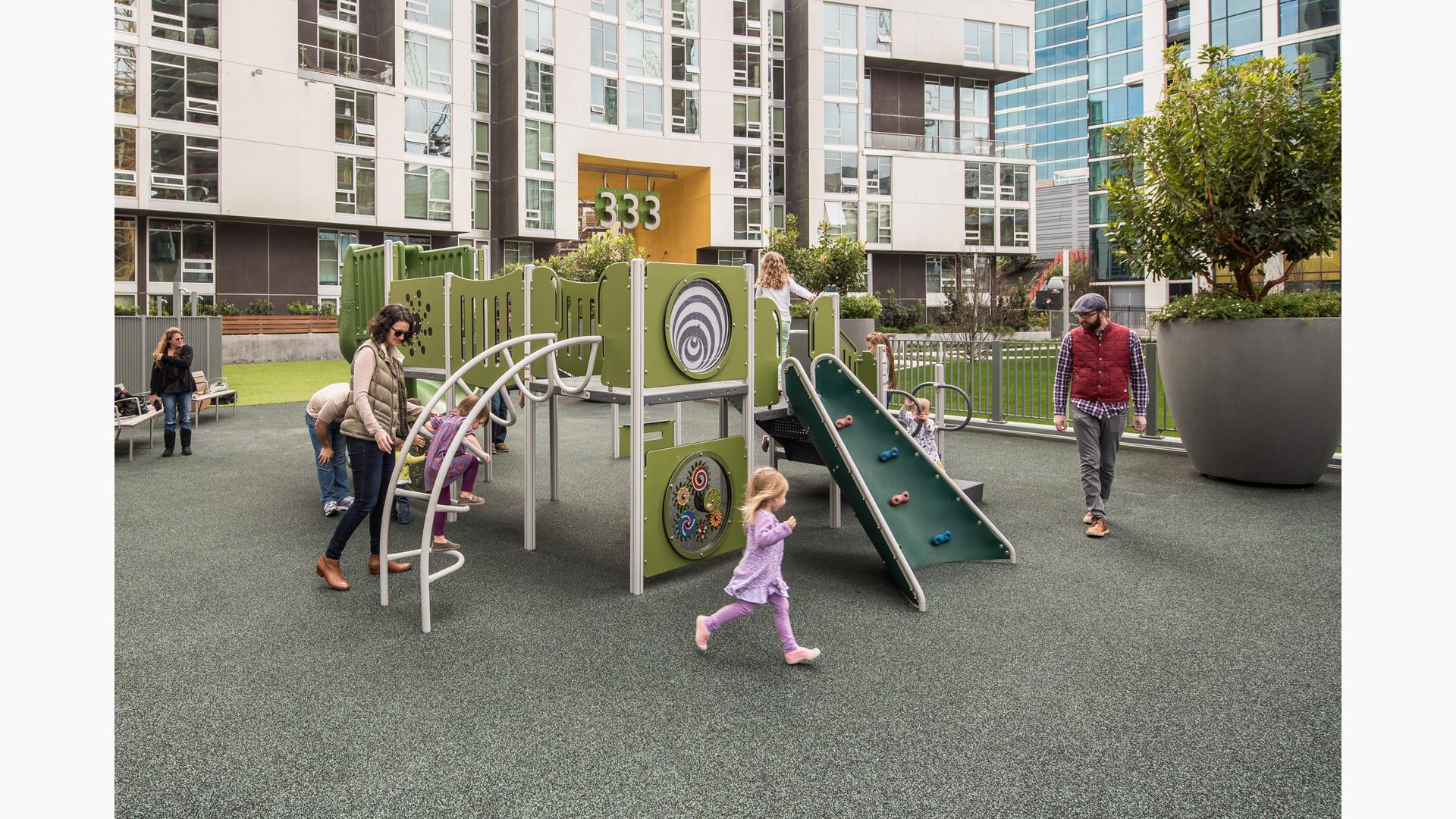 Parents watch as their toddlers run and play on the PlayShaper play structure on the rooftop of their apartment complex.