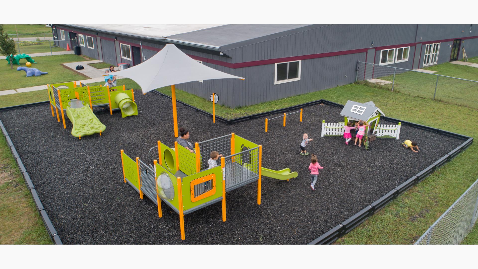 Elevated view of a play area set next to a grey building. Children play on three separate play structures colored in lime green and yellow.