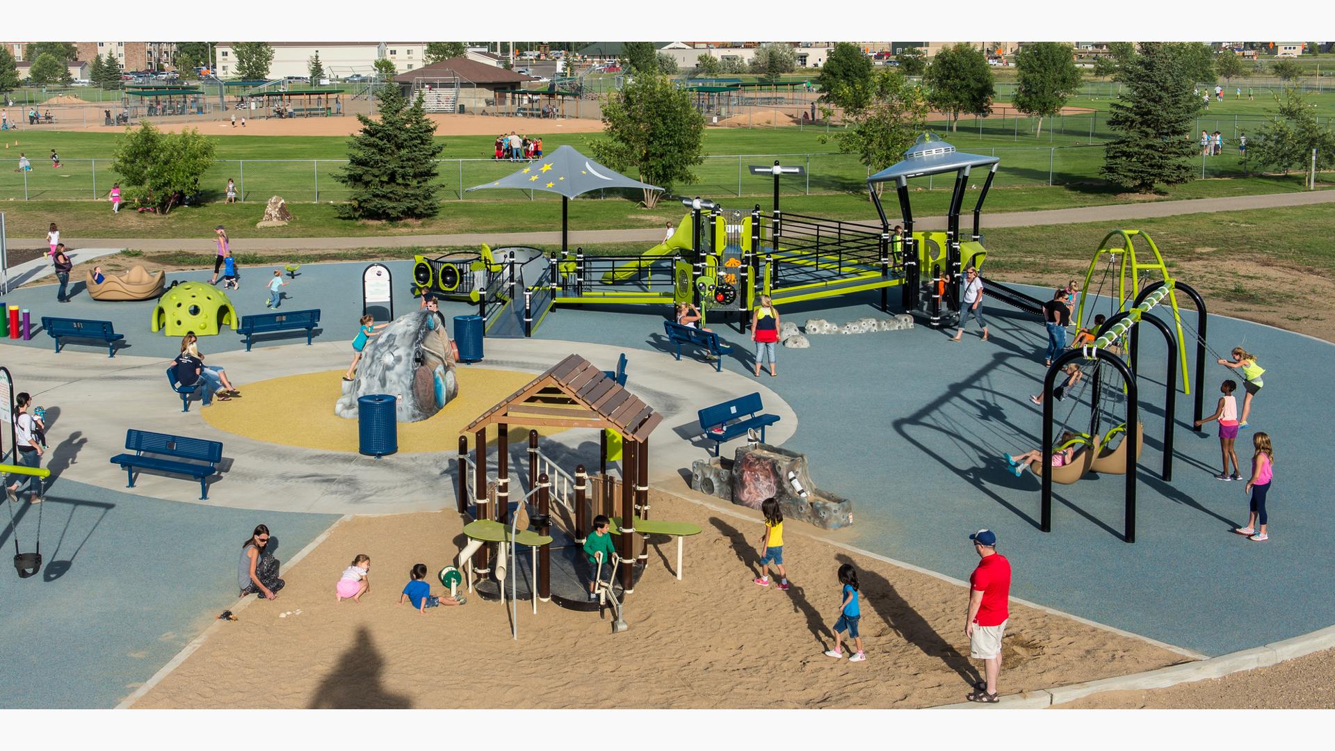 Children and families play on a sunny day in this alien-themed park. Featuring a UFO roof and a solar system sculpture in the middle of the playground.