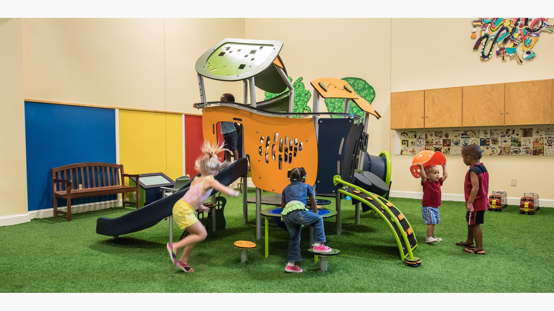 At a indoor play area young children play around a house like play structure with slides, climbers, and tunnels.