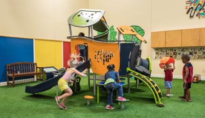 At a indoor play area young children play around a house like play structure with slides, climbers, and tunnels.