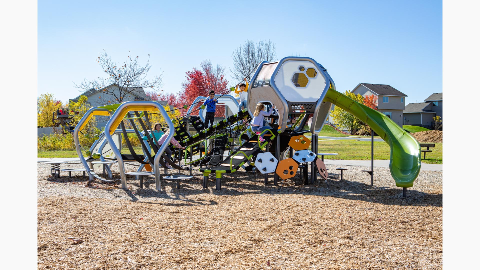 On a sunny day children bundled up in coats playing on futuristic playground structure with lots of hexagonal shapes. Neighborhood homes line the background.