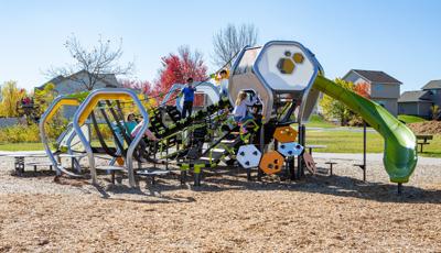 On a sunny day children bundled up in coats playing on futuristic playground structure with lots of hexagonal shapes. Neighborhood homes line the background.