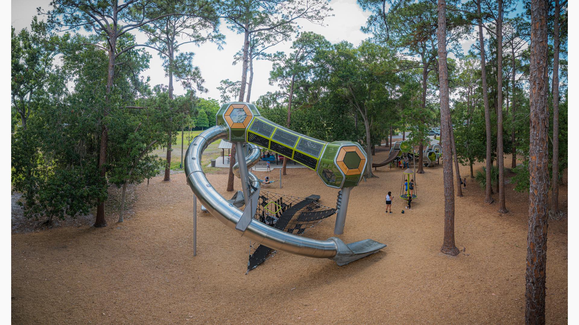 Two tower structures with hexagonal play pods are connected by a enclosed crawl tunnel and tubular stainless steel slides. Tall trees surround the play area.  