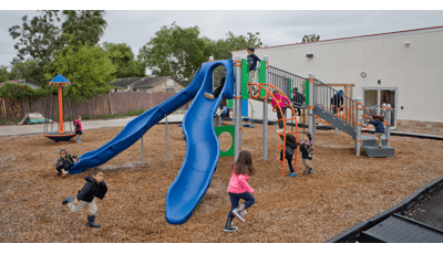 On a cloudy day children at Promesa College Prep play on the playground. A boy is about to ride down the slide, while another runs at full speed across the playground. Three children clamor onto the Loop Arch.