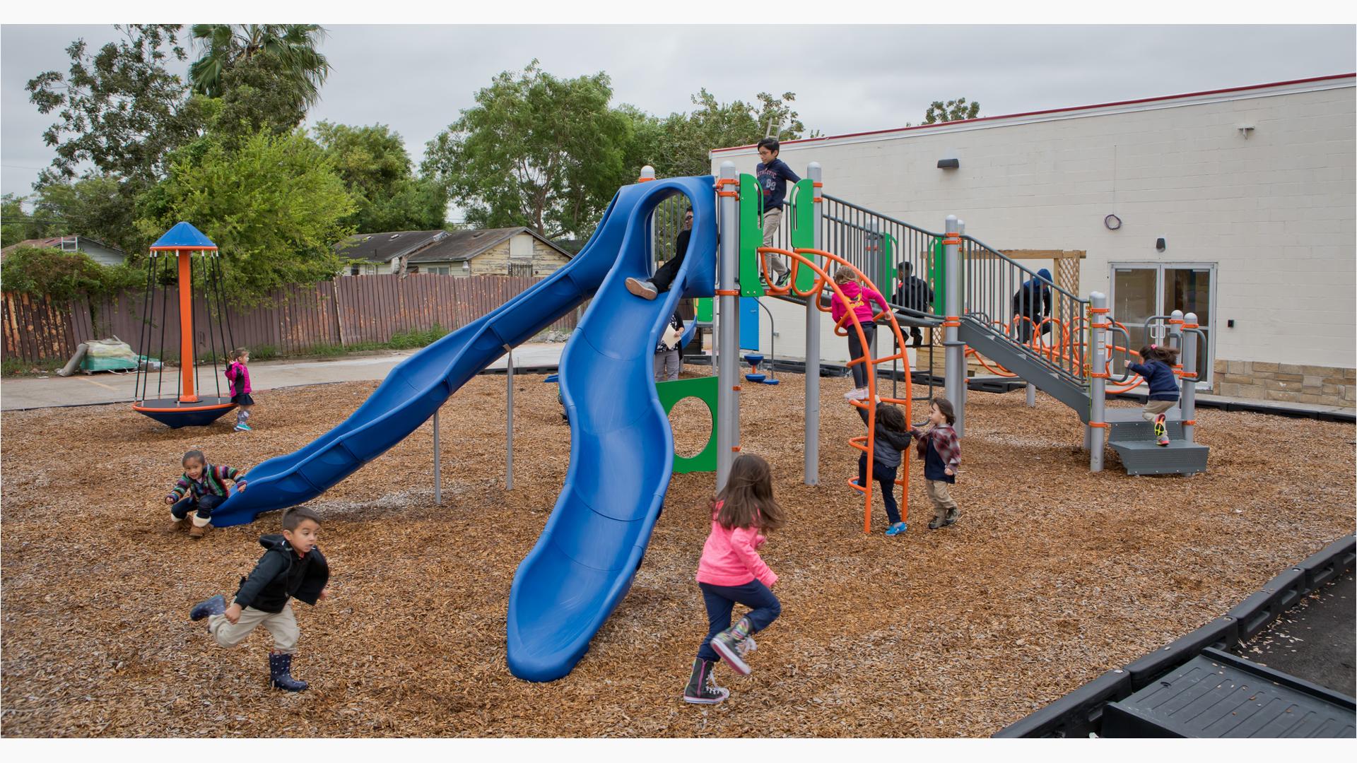 On a cloudy day children at Promesa College Prep play on the playground. A boy is about to ride down the slide, while another runs at full speed across the playground. Three children clamor onto the Loop Arch.