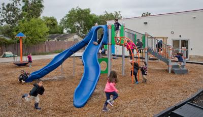 On a cloudy day children at Promesa College Prep play on the playground. A boy is about to ride down the slide, while another runs at full speed across the playground. Three children clamor onto the Loop Arch.