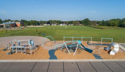 Full elevated view of a school playground area with two large separate play structures, swing sets, and a separate waved metal climber set on the edge of a open grass field with the school building off in the distance. 