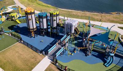 Elevated view of a large playground area set next to a pond. With a futuristic design four playground towers interconnected sit next to an additional play structure made of accessible ramps and three triangular shade sails overhead.