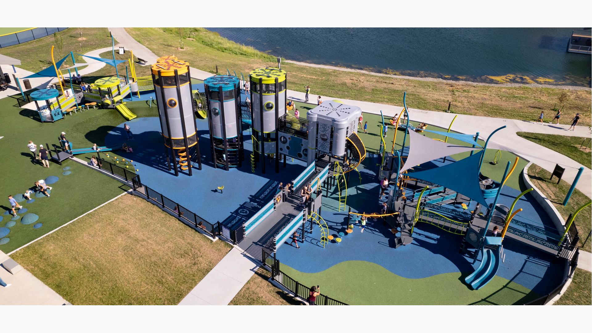 Elevated view of a large playground area set next to a pond. With a futuristic design four playground towers interconnected sit next to an additional play structure made of accessible ramps and three triangular shade sails overhead.