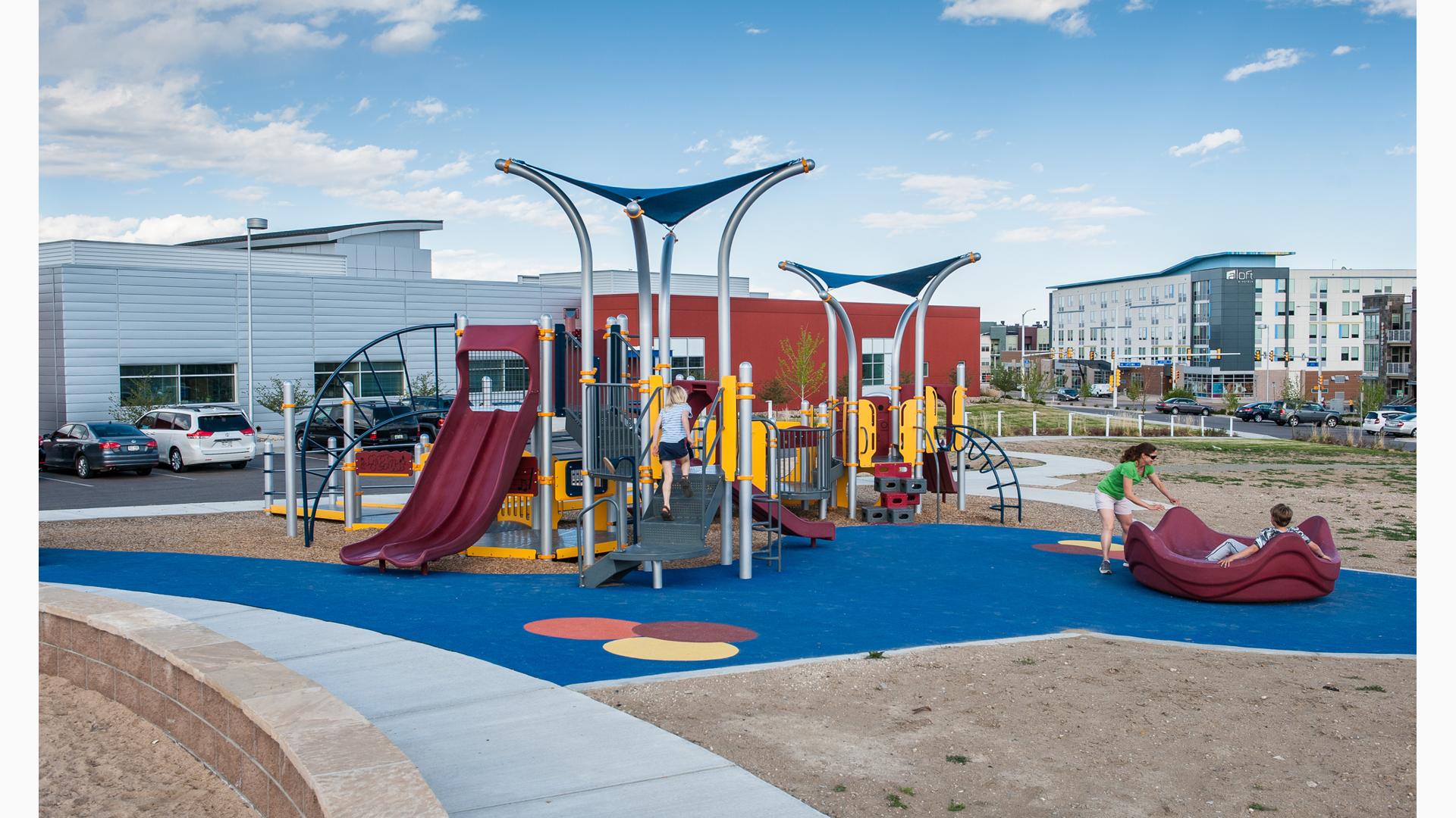 Girl walking up steps of playground in front of red and silver building white mother is pushing a boy in the merry-go-round. Surfacing is blue with sand on the outside. 