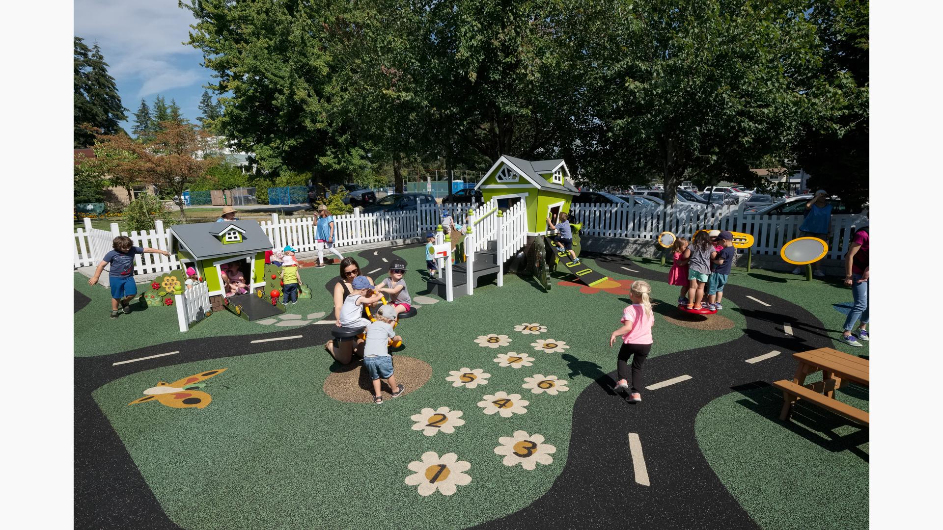 A white picket fence surrounds a play area with two play structure designed like small houses. The play area surfacing is designed with a winding street road, butterflies, and flowers.