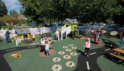 A white picket fence surrounds a play area with two play structure designed like small houses. The play area surfacing is designed with a winding street road, butterflies, and flowers.