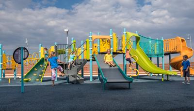 Boy in blue t-shirt watches as girl rides down Roller slide and two other children climb the stairs to the top of the play structure.