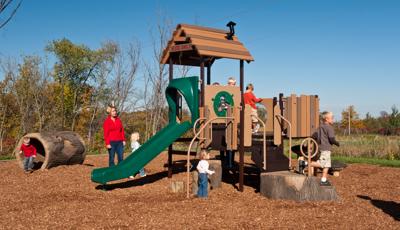 Mom watches children play on nature-inspired PlayShaper play structure on a sunny day.