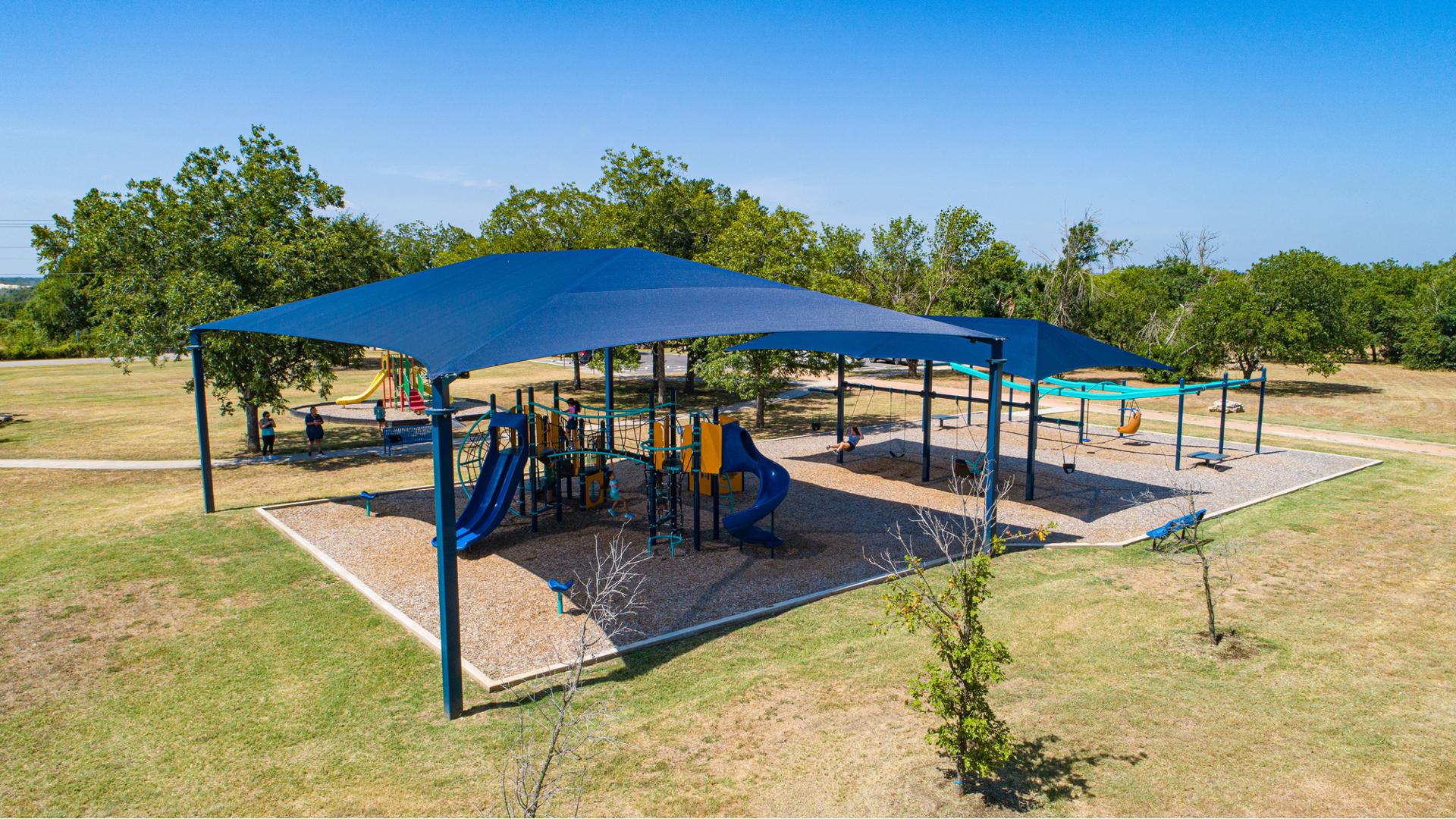 Elevated view of a large blue shade covering a playground structures with a secondary smaller shade right next to it covering a bay of swings.