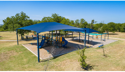 Elevated view of a large blue shade covering a playground structures with a secondary smaller shade right next to it covering a bay of swings.