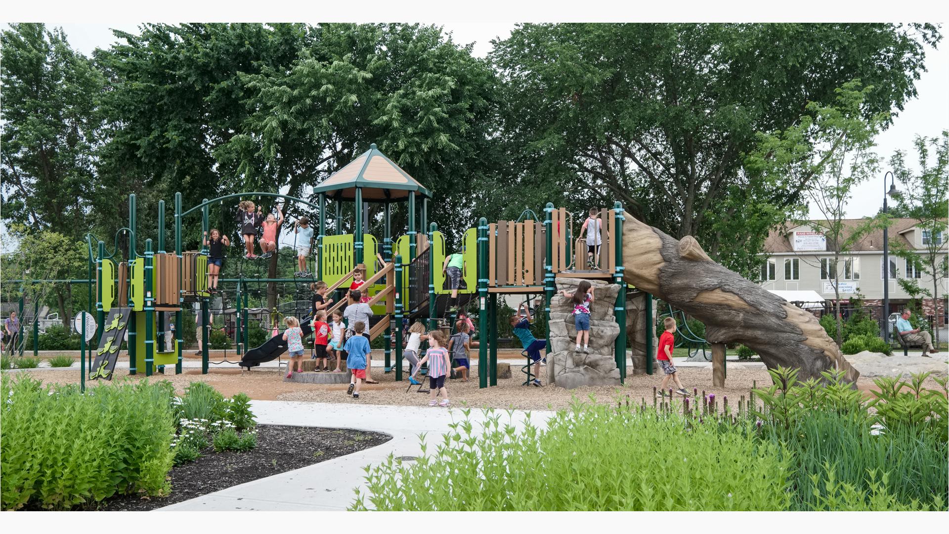 Nature inspired playground featuring log slide and fake rock climbing walls with young kids playing and climbing with tall trees and growing gardens surrounding the play space.