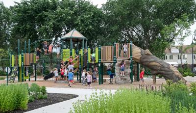 Nature inspired playground featuring log slide and fake rock climbing walls with young kids playing and climbing with tall trees and growing gardens surrounding the play space.