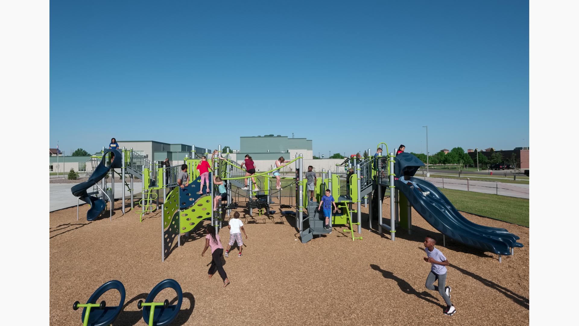 Children of Willow Park Elementary playing on new play structure. A large group climb on the Cascade climber. Two girls walk across net bridge. The line for the double slide is 3 kids deep as a boy runs across playground.