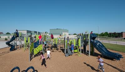 Children of Willow Park Elementary playing on new play structure. A large group climb on the Cascade climber. Two girls walk across net bridge. The line for the double slide is 3 kids deep as a boy runs across playground.