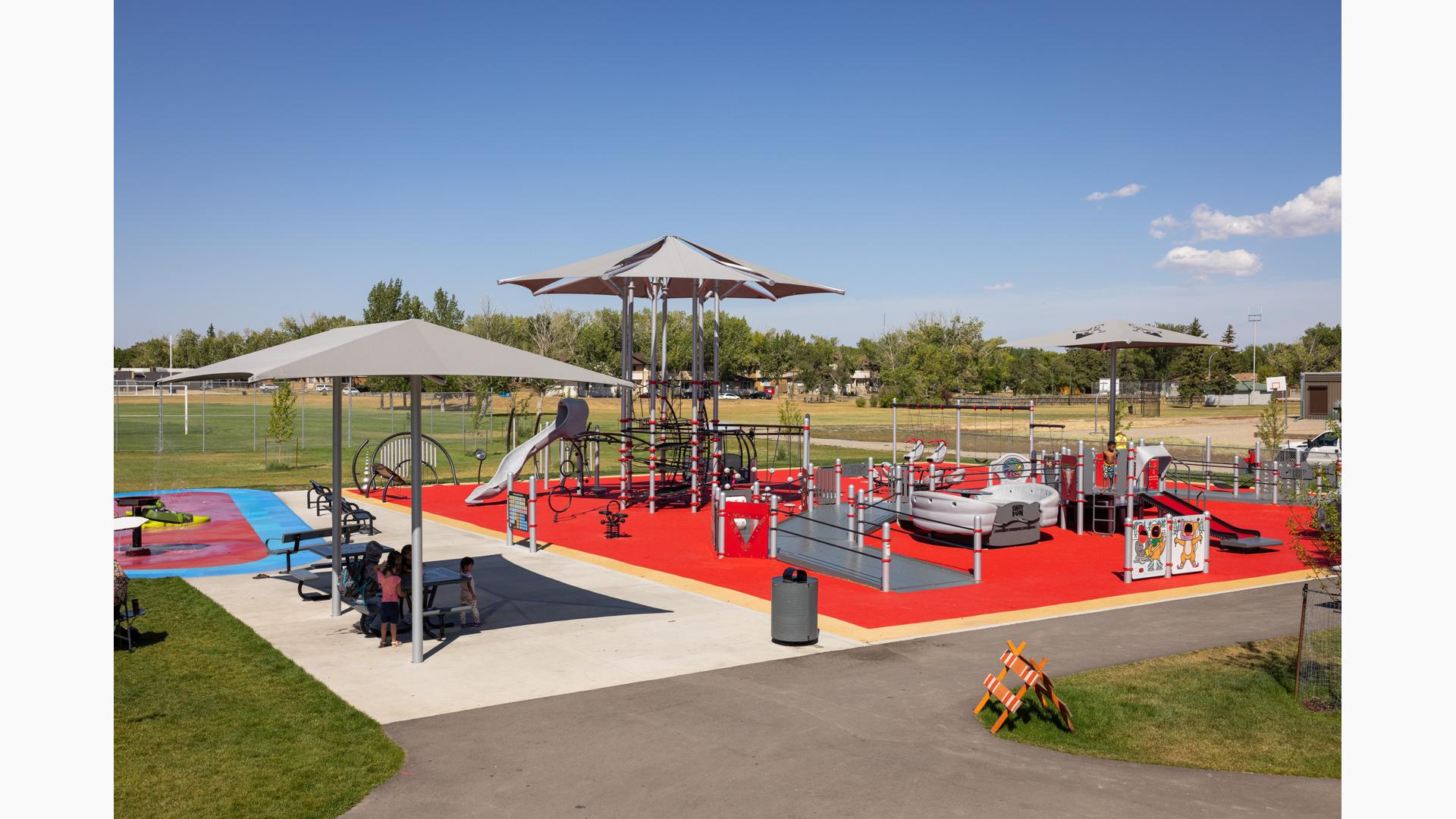 Children play on a large red and gray accented inclusive playground and parents sit at a picnic table of to the side under a shade.