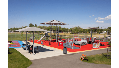 Children play on a large red and gray accented inclusive playground and parents sit at a picnic table of to the side under a shade.