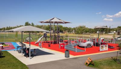 Children play on a large red and gray accented inclusive playground and parents sit at a picnic table of to the side under a shade.