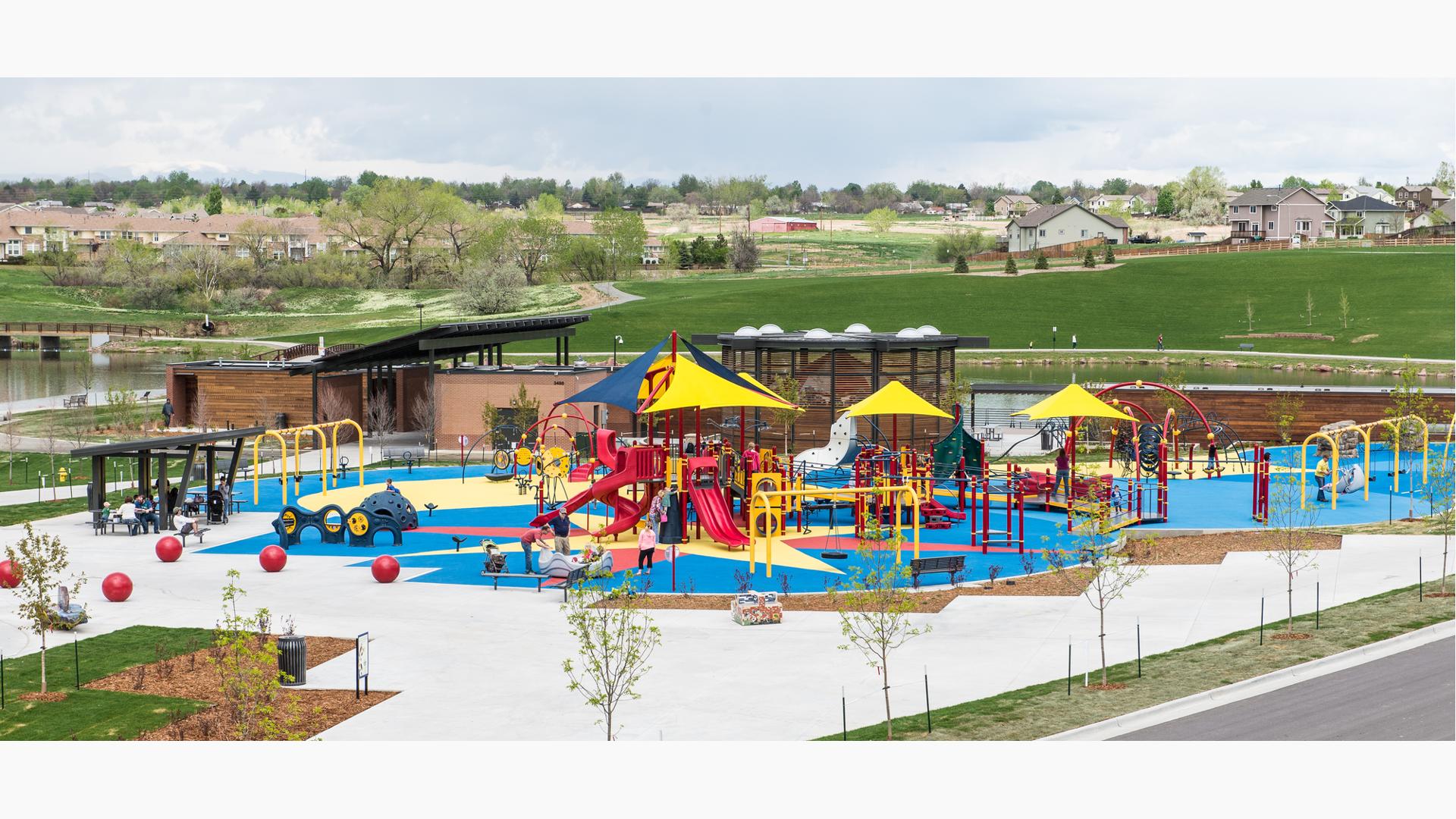 Full view of a park inclusive playground next to a pond with ramps, slides, swings, climbers, and overhead shade accessible to all ages.