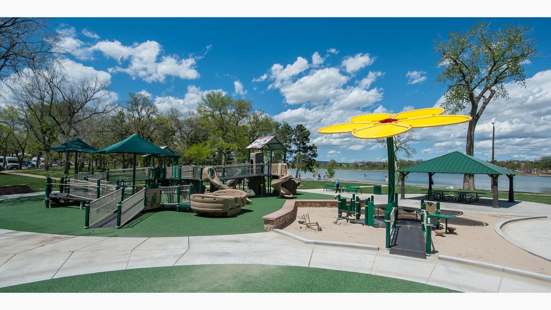 Playground structure with accessible ramps in tan and brown.  Play space also includes a yellow flower shaped shade structure with a lake in the background. 