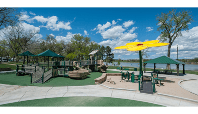 Playground structure with accessible ramps in tan and brown.  Play space also includes a yellow flower shaped shade structure with a lake in the background. 