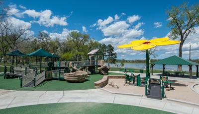 Playground structure with accessible ramps in tan and brown.  Play space also includes a yellow flower shaped shade structure with a lake in the background. 