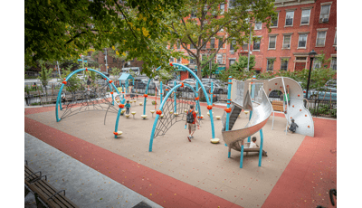 Blue arched playground with kids climbing on nets with a stainless steel slide located in a city park next to buildings with windows. 