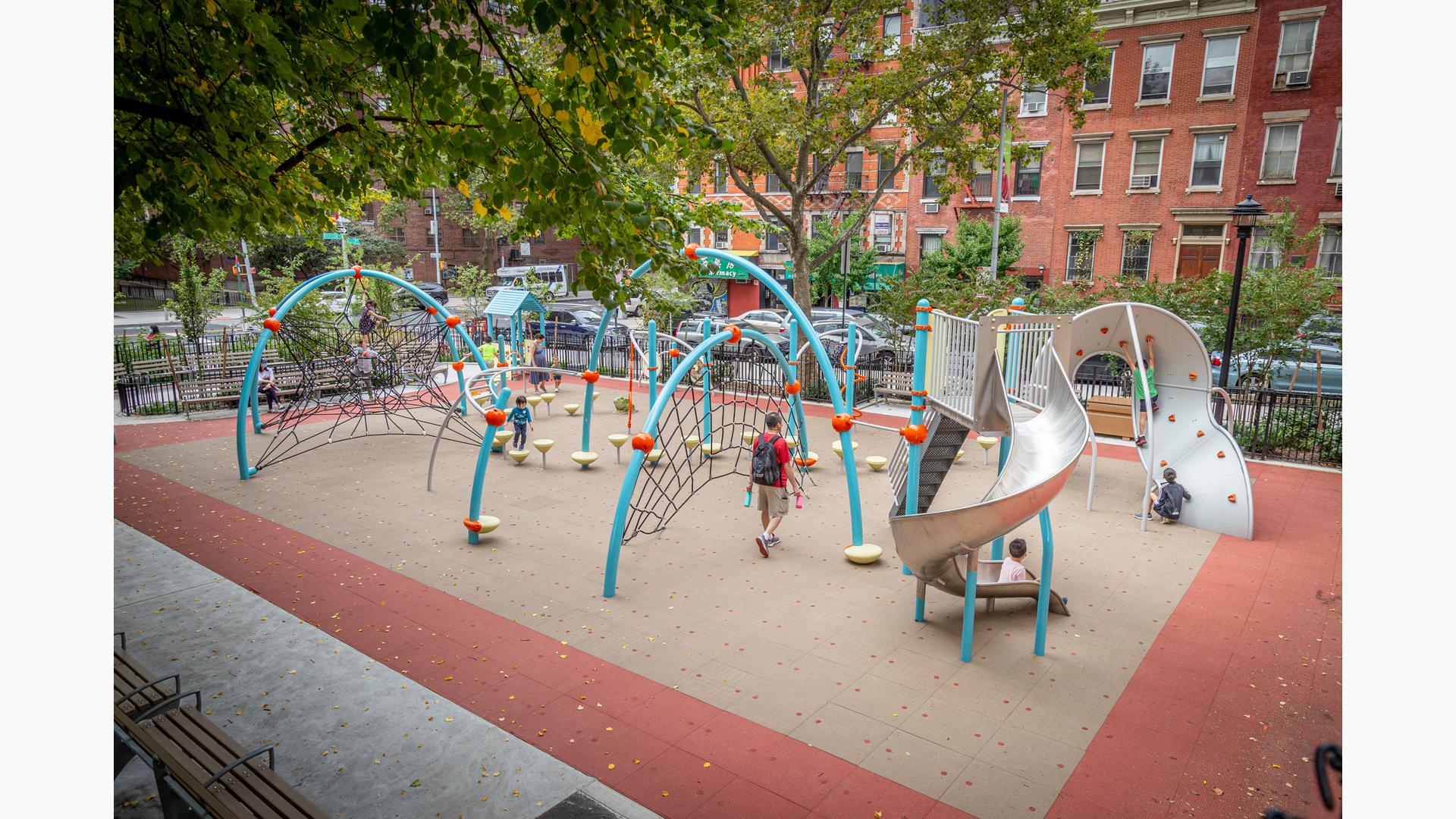 Blue arched playground with kids climbing on nets with a stainless steel slide located in a city park next to buildings with windows. 