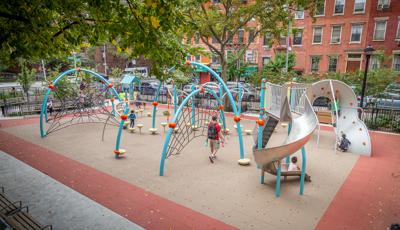 Blue arched playground with kids climbing on nets with a stainless steel slide located in a city park next to buildings with windows. 