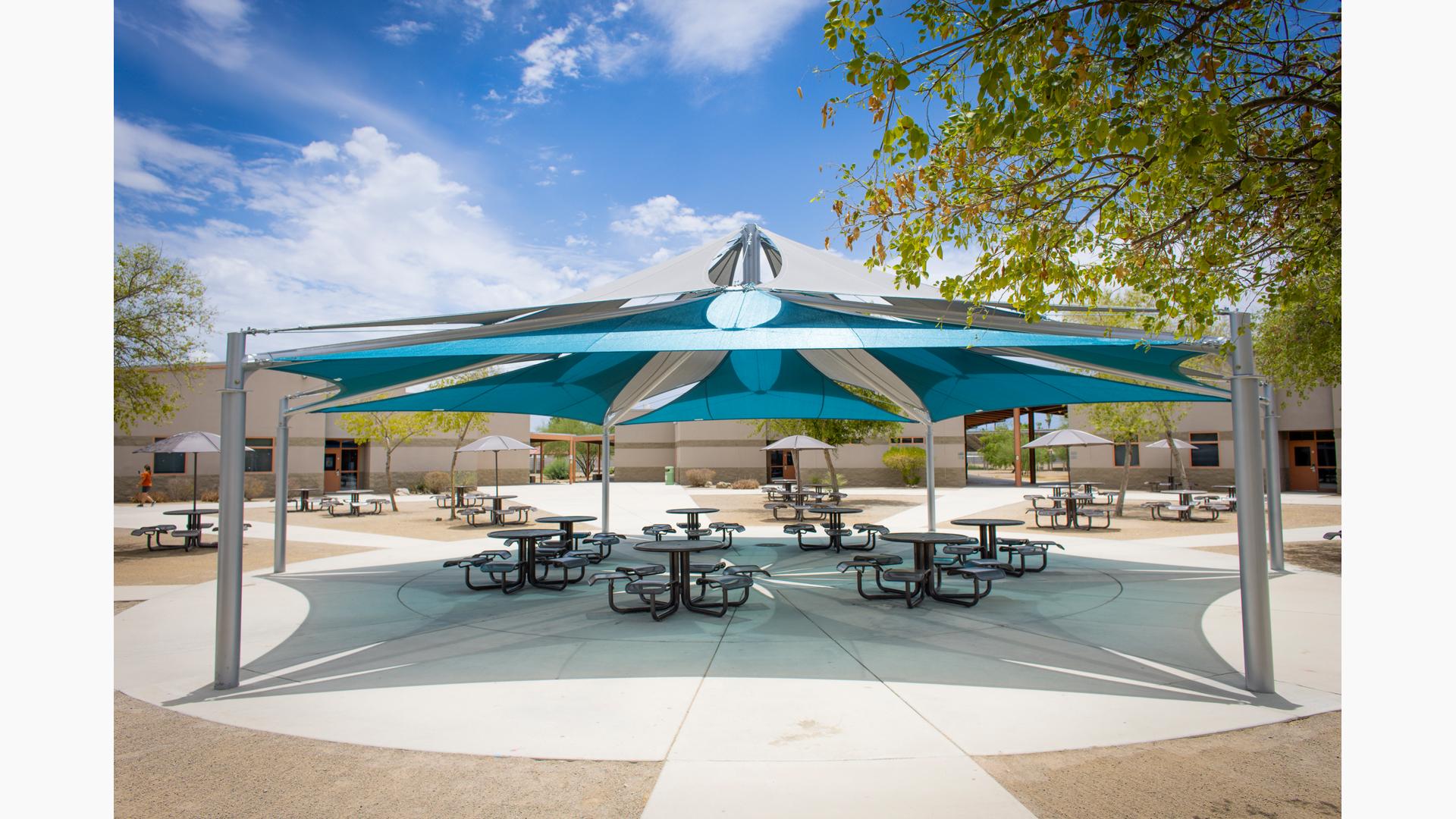 A large hexagonal multi overlapping shade system stands over a circular concreated courtyard scattered with many picnic tables. Three separate buildings stand in the background with leading walk ways to the open courtyard.