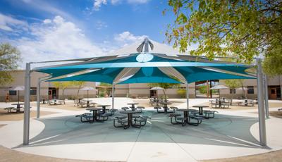 A large hexagonal multi overlapping shade system stands over a circular concreated courtyard scattered with many picnic tables. Three separate buildings stand in the background with leading walk ways to the open courtyard.